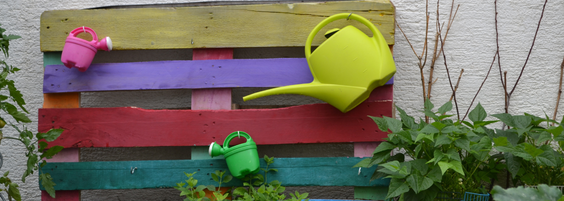 Farbige Holzpalette mit bunten Plastikgieskannen vor einem Gebüsch in der Natur draußen.